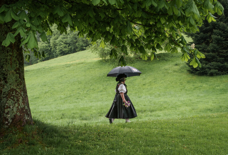 Das Bild zeigt eine Frau mit der Bollenhuttracht unter einem Schirm über eine Wiese laufen. Sie trägt den Bollenhut in schwarz, was den verheirateten Frauen zusteht.